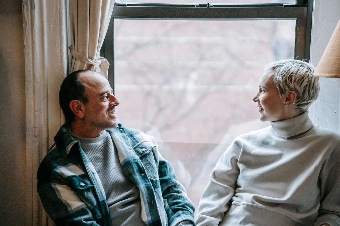 A couple sitting together by a window, looking at each other and smiling, sharing an unhurried moment