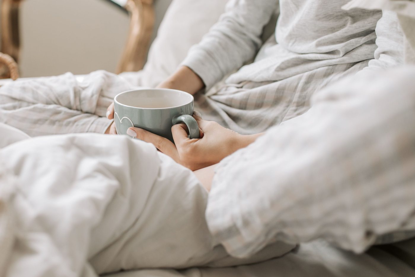 A person sitting in bed under a soft white duvet, holding a mug in both hands in quiet morning light