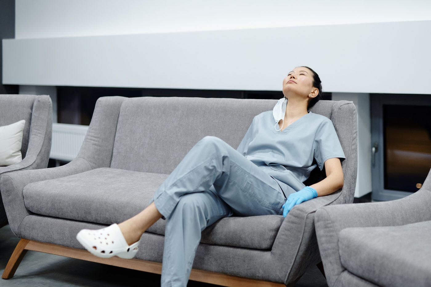 A healthcare worker in scrubs resting with eyes closed on a couch, mask pulled down, exhausted after a shift